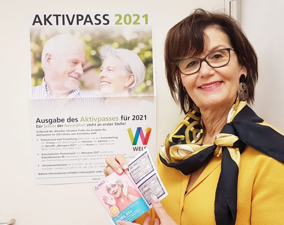 Stadträtin lächelt in Kamera mit dem aktuellen Aktivpass-Heft für das Jahr 2021. Im Hintergrund ein Plakat mit Richtlinien zur Ausgabe