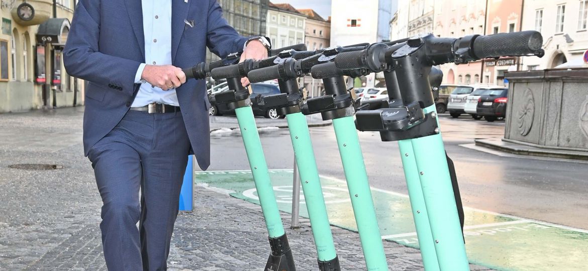 Vizebürgermeister Gerhard Kroiß steht hinter einer Gruppe von E-Scootern auf dem Stadtplatz, im Hintergrund ist der Ledererturm zu sehen