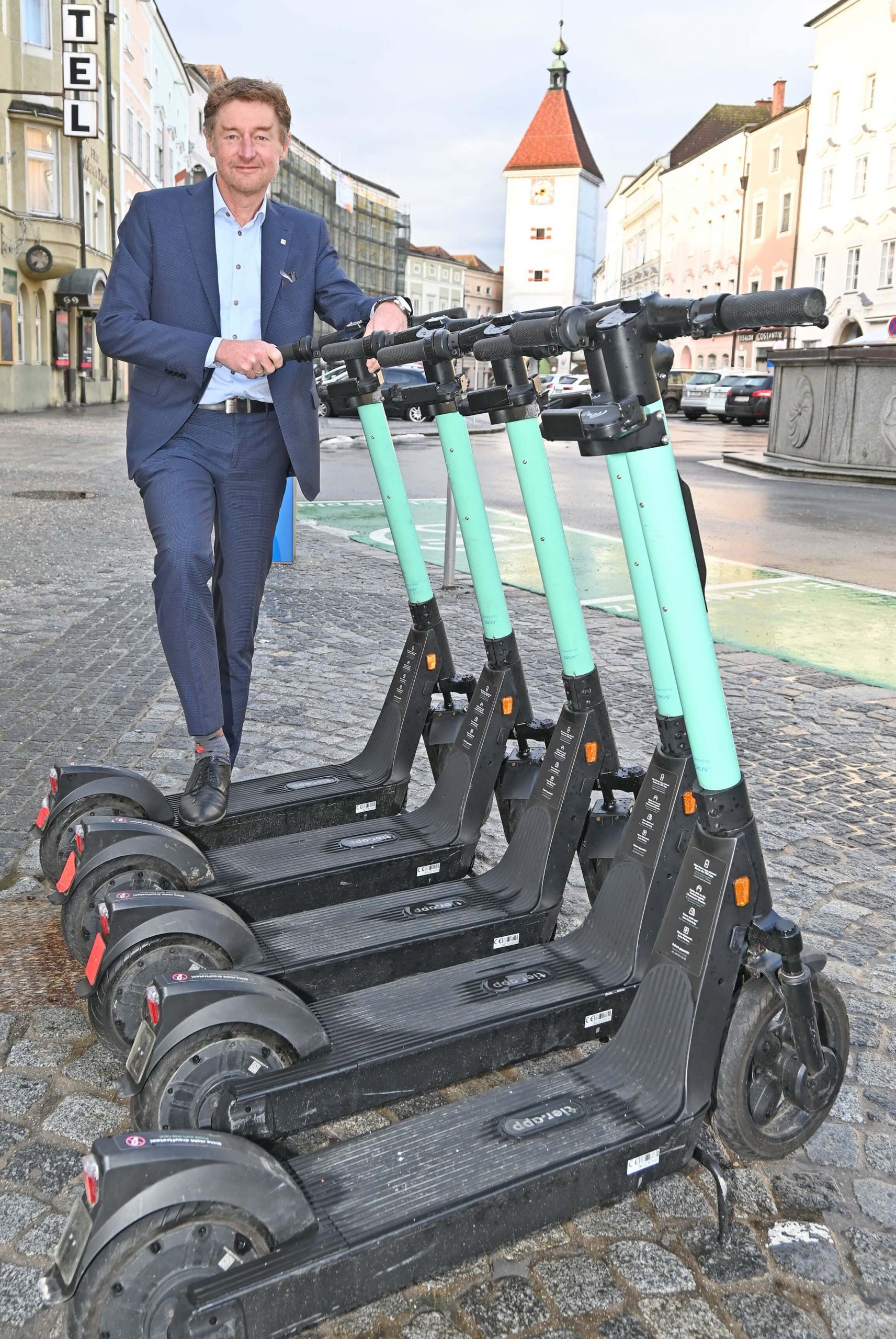Vizebürgermeister Gerhard Kroiß steht hinter einer Gruppe von E-Scootern auf dem Stadtplatz, im Hintergrund ist der Ledererturm zu sehen