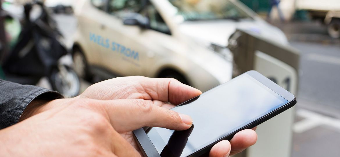 Eine Hand die auf ein Handy tippt und im Hintergrund ist ein E-Auto