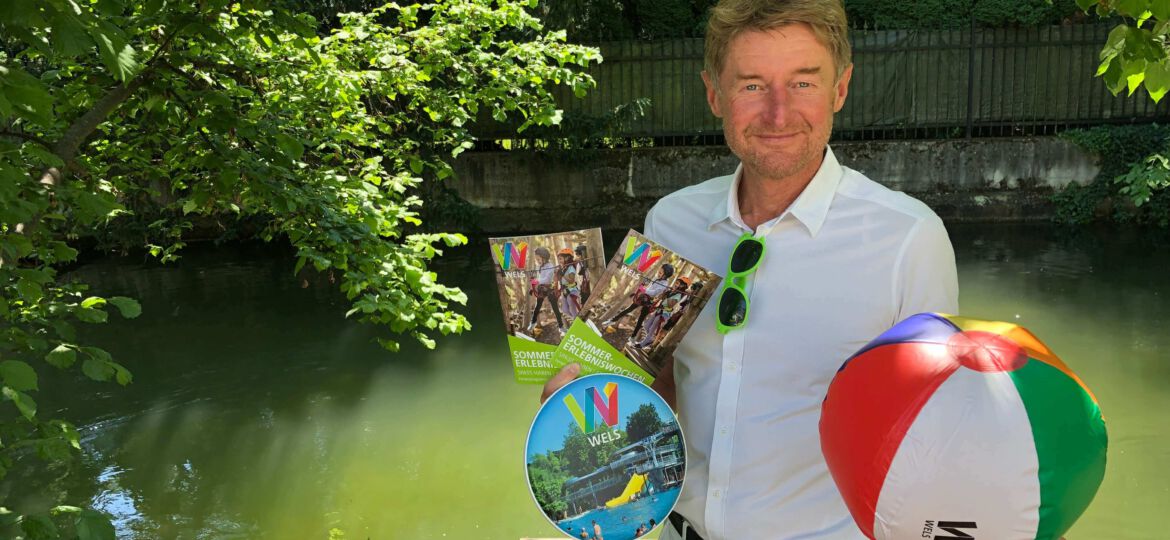 Vzbgm. Gerhard Kroiß mit einem Wasserball, Frisbee und dem diesjäjrigen Sommerferienprogramm auf einem Steg beim Welser Mühlbach