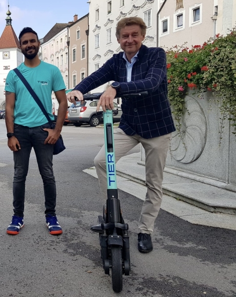 Herr Vizebürgermeister Kroiß steht mit einem E-Scooter und einem Mitarbeiter der Firma Tier neben dem Brunnen vor dem Rathaus mit Blick auf den Ledererturm