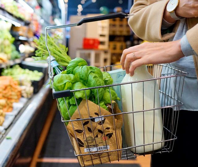 Einkaufskörbchen mit Lebensmittel, gehalten von einer Frau, im Hintergrund ein Supermarktregal