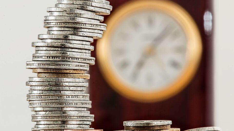 Stapel Gold- und Silbermünzen im Vordergrund, eine Wanduhr verschwommen im Hintergrund